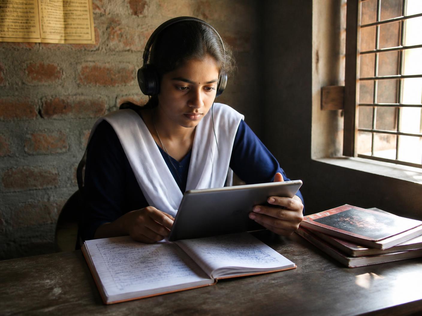 A teenage student studying with a tablet and notebook at a desk beside a sunlit window at home